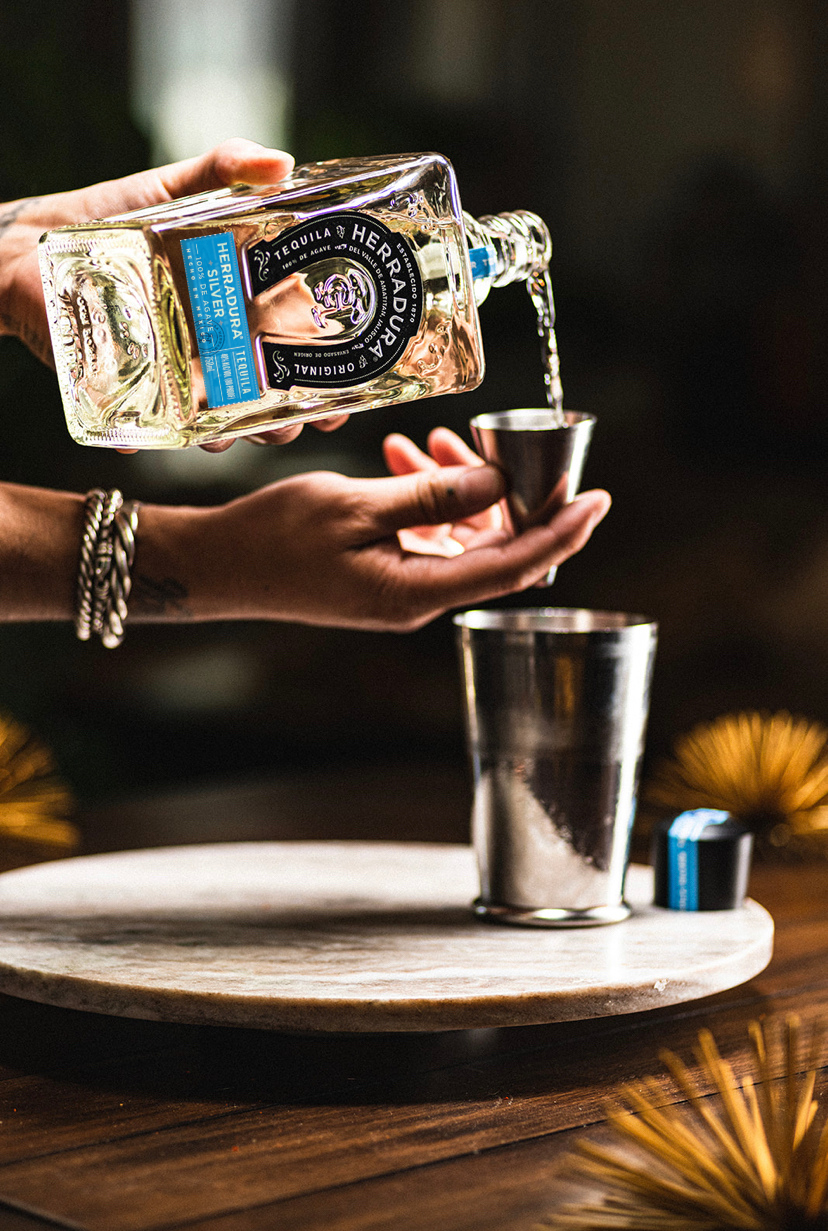 A tattooed hand pours tequila into a cocktail shaker