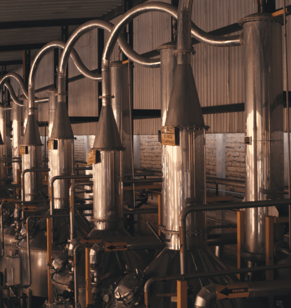 Industrial distillation columns with interconnected metal pipes inside a manufacturing facility, likely used for producing spirits or other distilled products