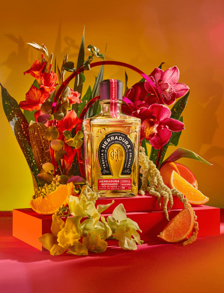 An image of a bottle of Herradura Reposado on a platform surrounded by florals and citrus in front of a golden toned background.