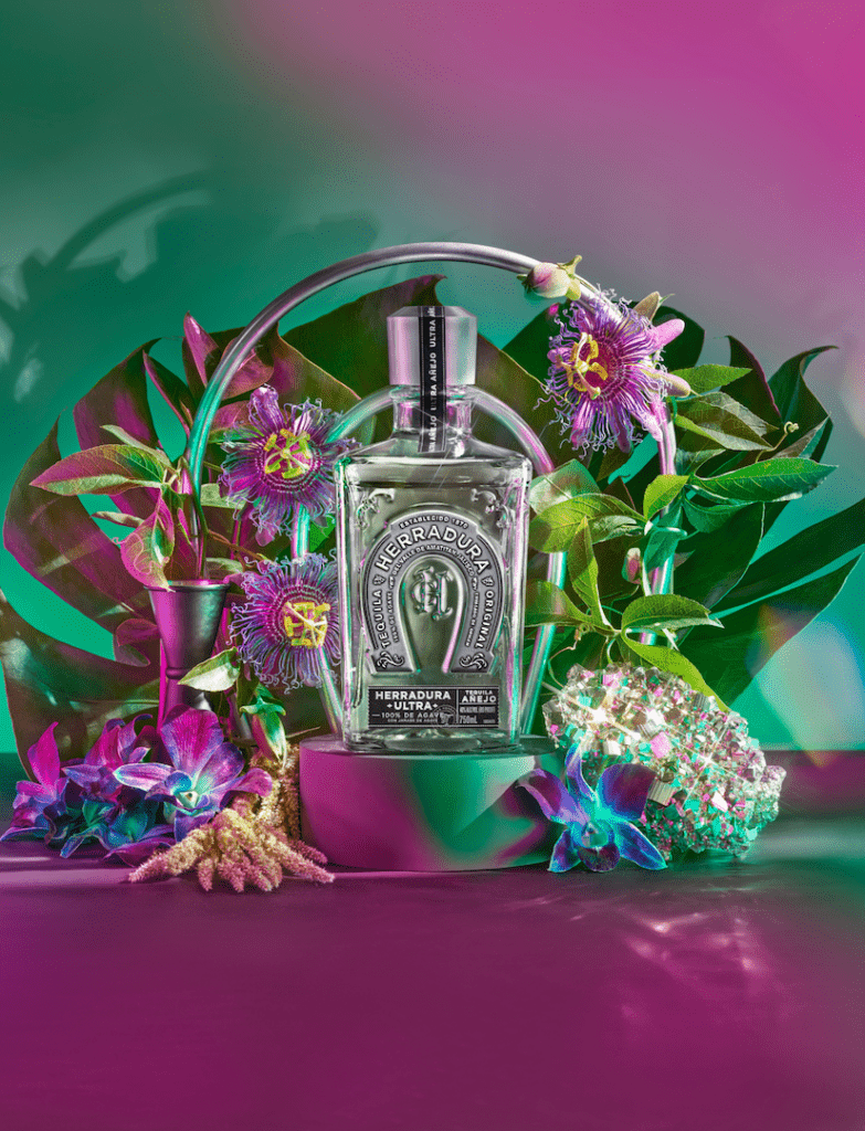 An image of a bottle of Herradura Ultra on a platforms surrounded by florals in front of a green and pink ombre background